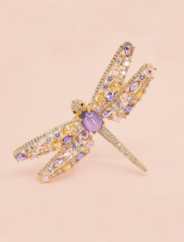 Broche dorado en forma de libélula con cristales brillantes y piedra central lila.